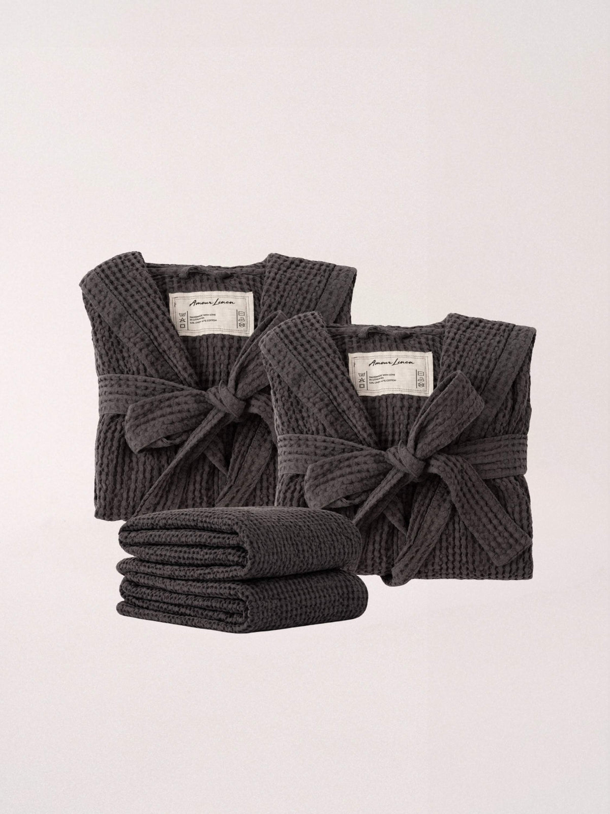 Three dark gray waffle-knit robes in different sizes, with two standing upright and one folded in front, on a plain light background. Each robe has a tie belt and a small white brand label.